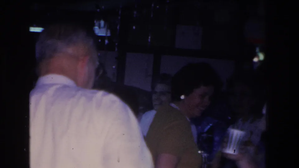 A man in a white shirt with his back to the camera occupies the foreground while other adult guests socialize behind him in a dimly lit interior, from a 1966 8mm home movie from Hoboken, New Jersey.