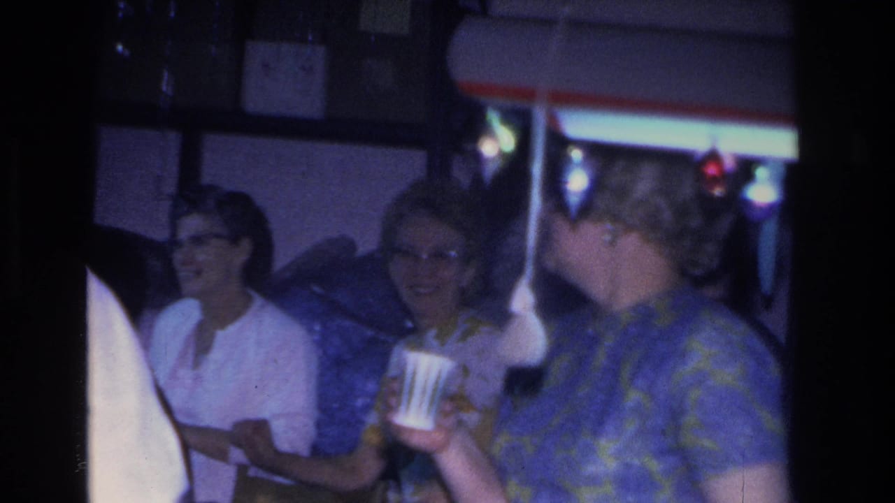 Candid 8mm home-movie frame from a 1966 Christmas party in Hoboken, New Jersey; older women socializing under colored string lights, one holding a paper cup.