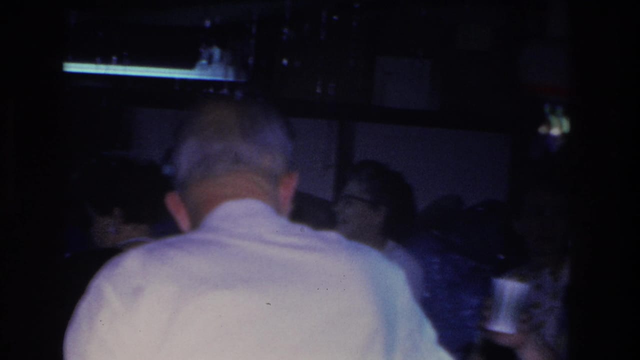 Man in white shirt seen from behind dominates the frame; blurred figures visible in background including a woman with glasses; dim interior lighting.