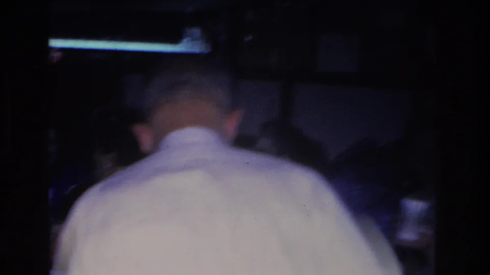 8mm frame showing an older man from behind in a white shirt at an indoor 1966 holiday gathering; the lower frame is underexposed and background subjects are out of focus.