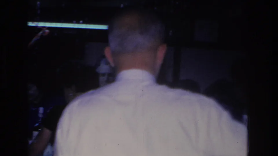 8mm frame at a 1966 indoor Christmas party; the same man in white shirt is again seen from behind, with a darker figure to his right and what may be tabletop items visible in the lower right corner.