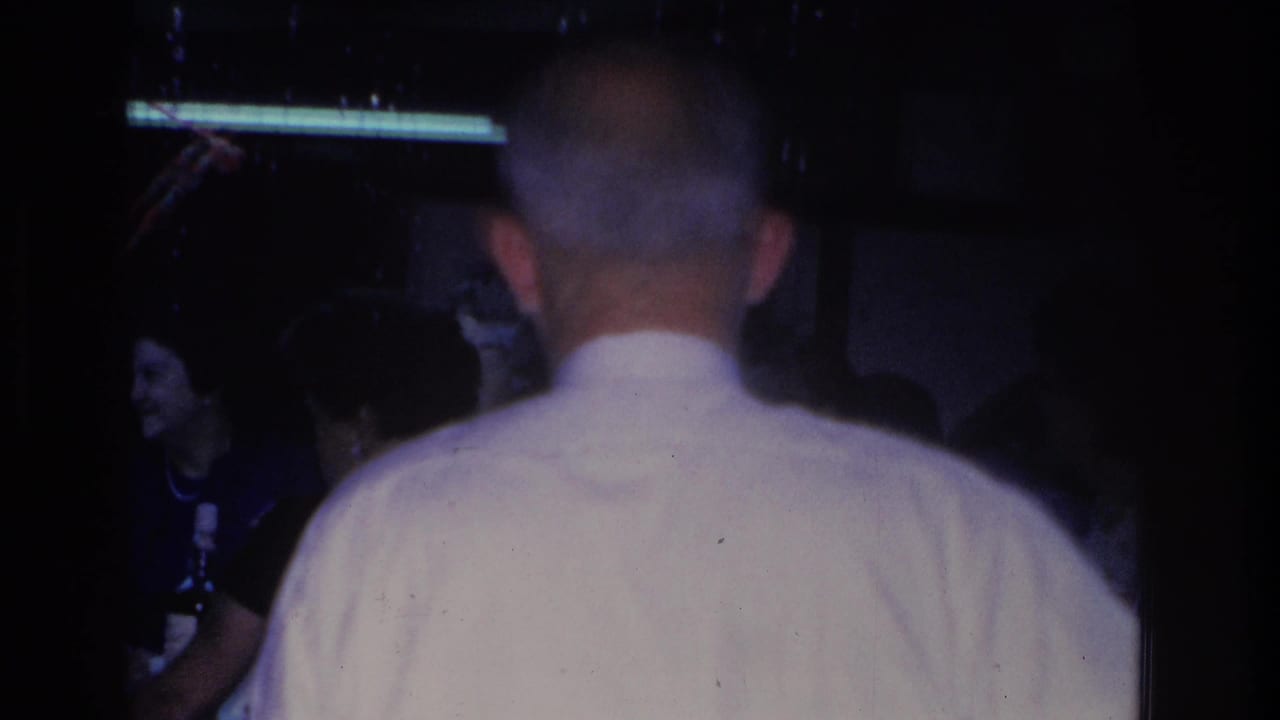 8mm home movie frame, circa 1966, showing the back of a man in a white shirt at an indoor social gathering in Hoboken, New Jersey; a smiling woman is partially visible at left.