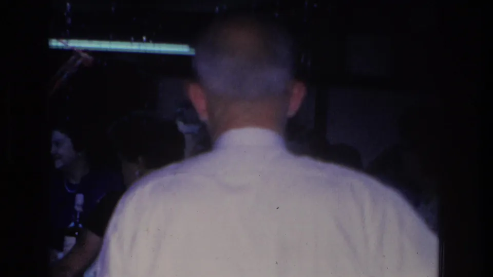 8mm home movie frame, circa 1966, showing the back of a man in a white shirt at an indoor social gathering in Hoboken, New Jersey; a smiling woman is partially visible at left.