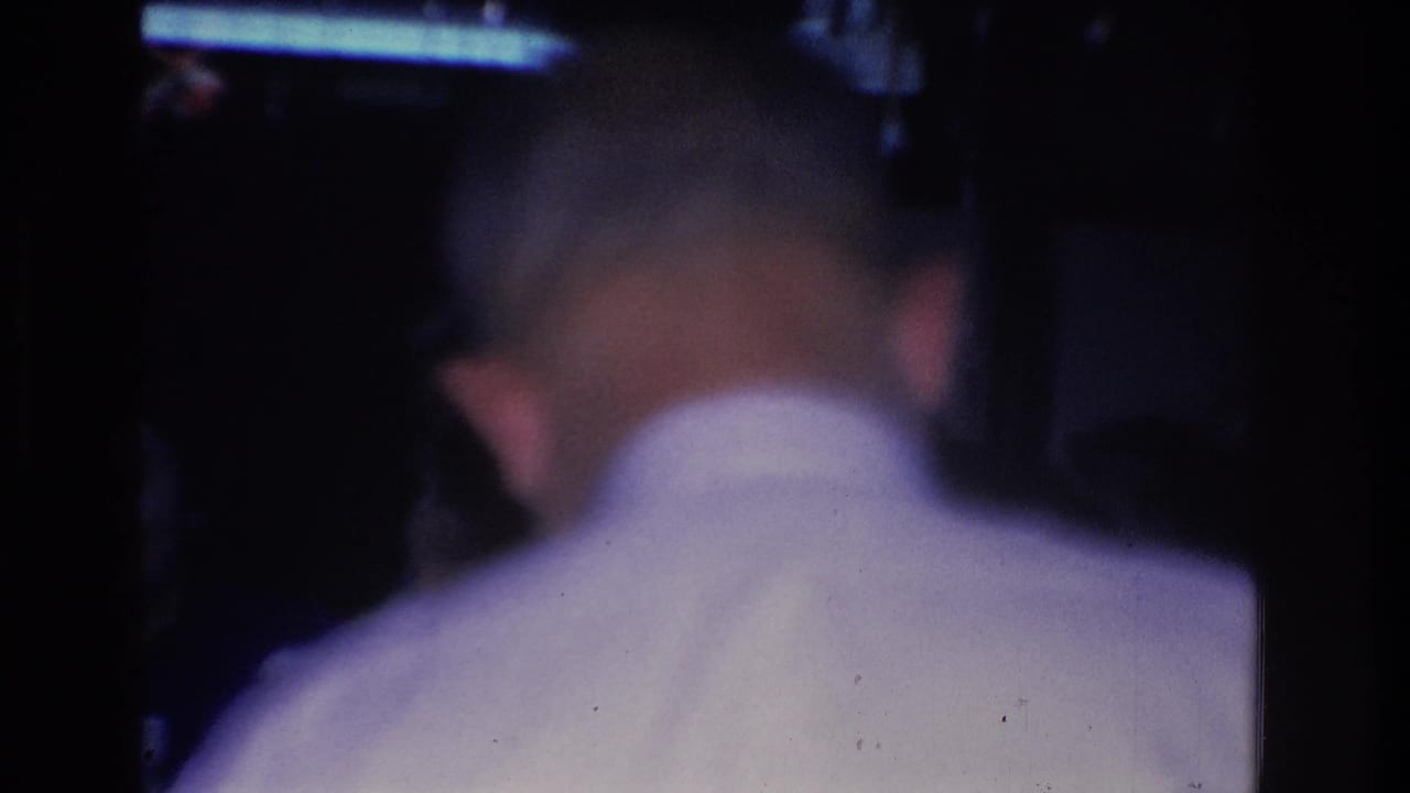 Close view of a man's back in a white collared shirt at a dark indoor gathering; a fluorescent light fixture is visible at upper right.