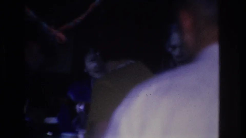 8mm home movie frame, circa 1966, a woman's face partially visible amid a very dark interior gathering; man in white shirt remains at right frame edge; indoor social event, Hoboken, New Jersey.