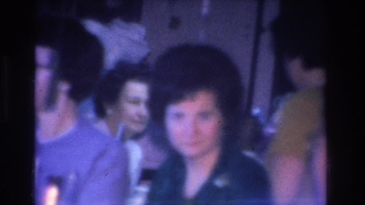 Group of women in 1960s clothing gathered indoors, blue-purple 8mm film cast, soft focus.
