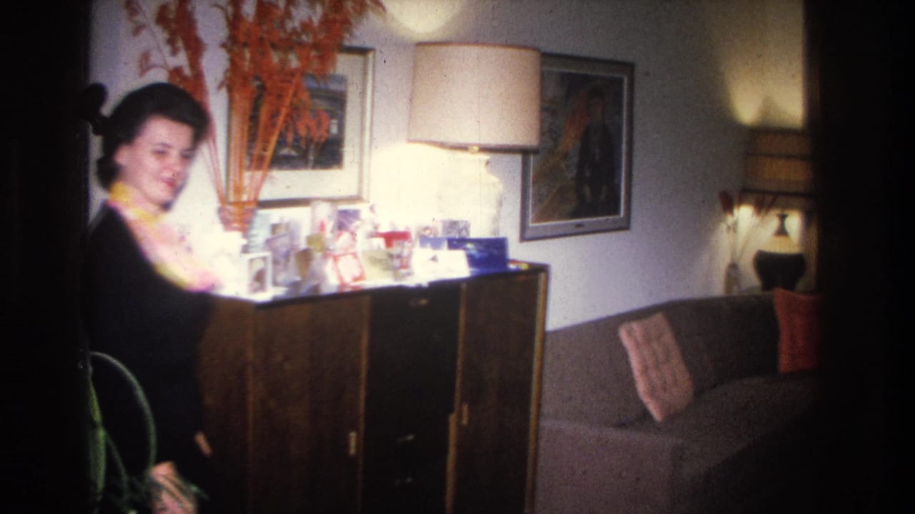 Woman in black dress with yellow scarf smiles toward camera in a 1966 Hoboken, NJ home, beside a dark wood sideboard bearing greeting cards and photos.