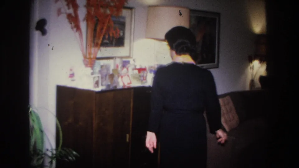 Back view of woman in black dress approaching a sideboard in a 1966 home interior, Hoboken, NJ; sofa, wall art, and floral arrangement visible.