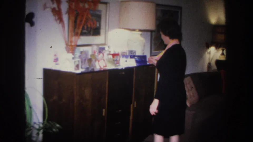 Side profile of woman in black dress reaching toward a mid-century sideboard in a 1966 Hoboken home interior; greeting cards and decorative items on cabinet top.