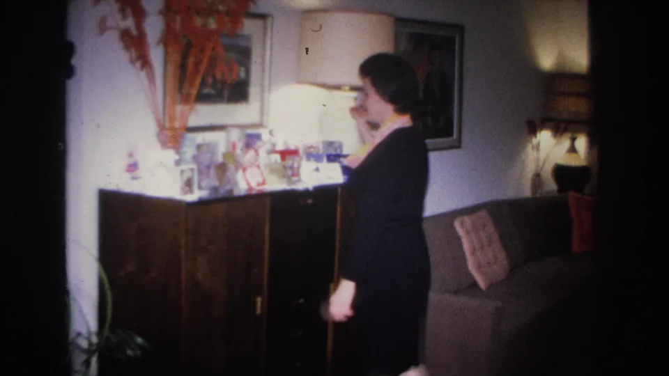 Woman in black dress and eyeglasses at a sideboard in a 1966 Hoboken home interior; greeting cards, small objects, floor lamp, and sofa visible.