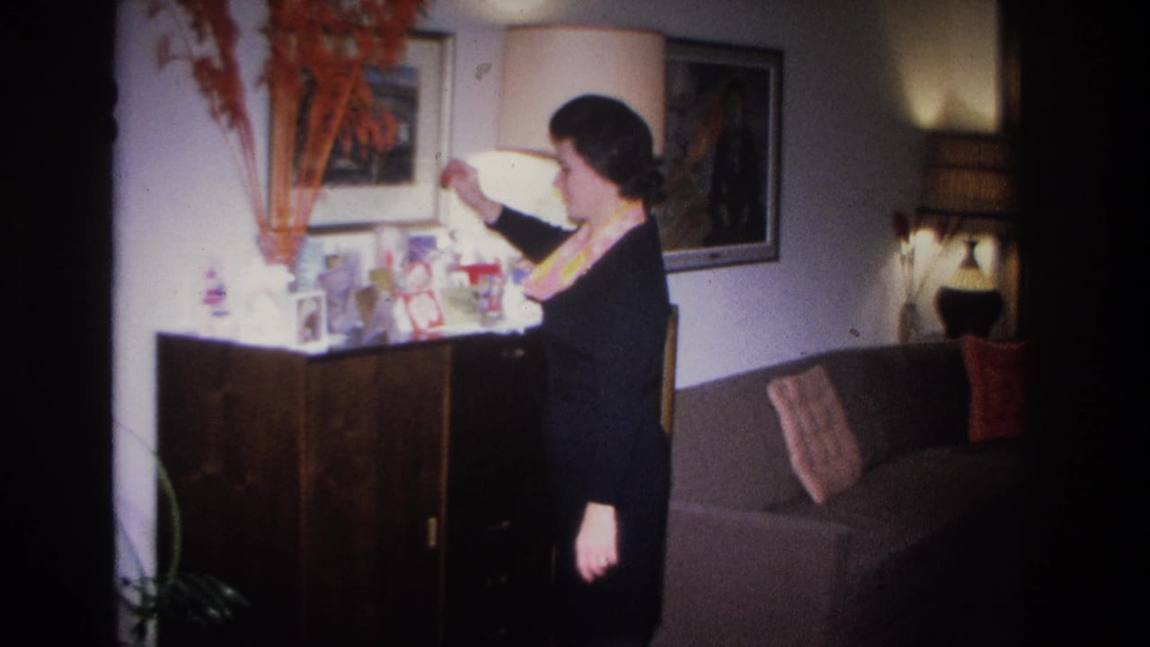 Woman in dark dress and yellow scarf tends to small objects on a mid-century sideboard; framed artwork and lamp visible; Hoboken, New Jersey, circa 1966.