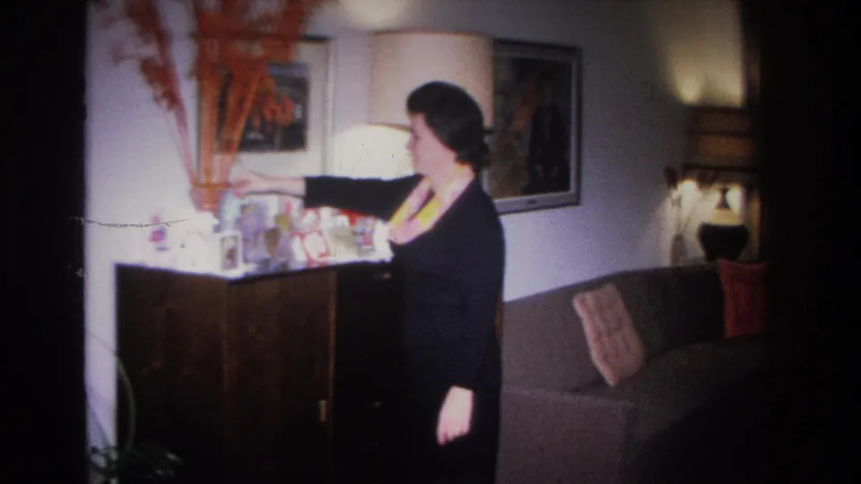 Woman in dark dress pauses beside an open sideboard in a mid-century American living room; table lamp and artwork on wall, Hoboken, New Jersey, circa 1966.