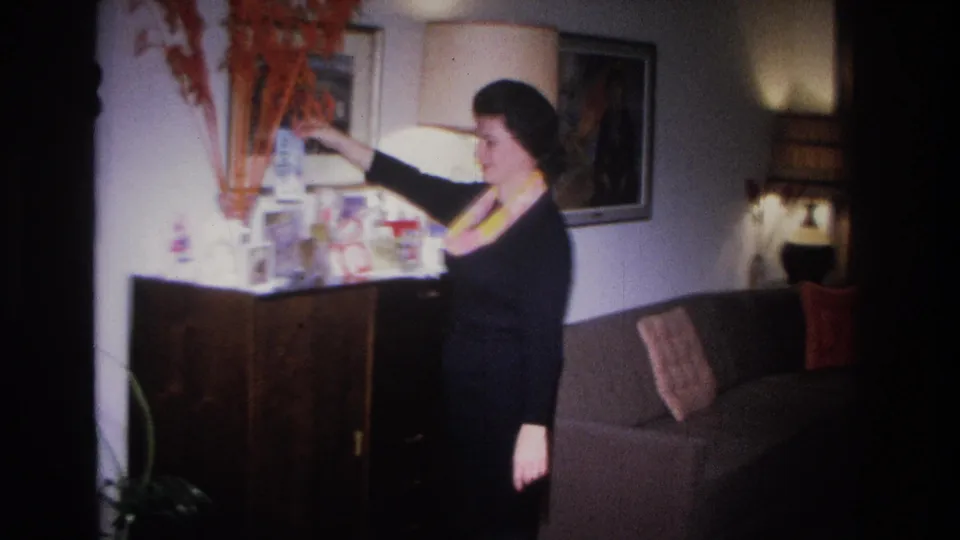 Woman reaching to adjust small objects on a mid-century sideboard in a 1960s living room; sofa with cushions and lamp visible behind her, circa 1966.
