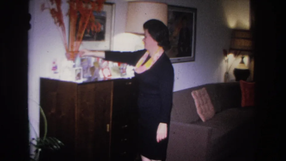 Woman in dark dress beside dark-wood sideboard in a mid-century living room; lamp glowing and wall art visible, Hoboken, New Jersey, circa 1966.
