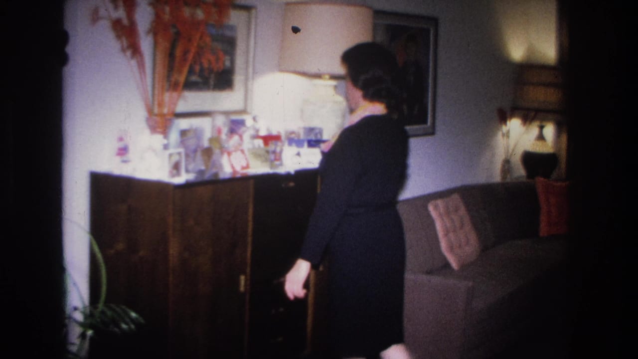 Woman in dark dress attending to items on a wood credenza in what appears to be a 1960s New Jersey living room; tufted sofa and bold framed wall art visible behind her.