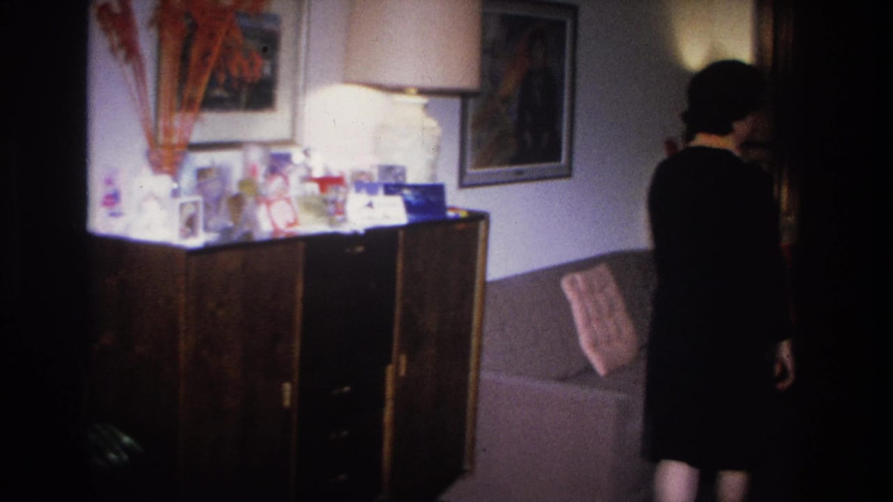Woman in dark dress near right edge of frame, viewed from behind, with wood sideboard and bold framed wall artwork visible.