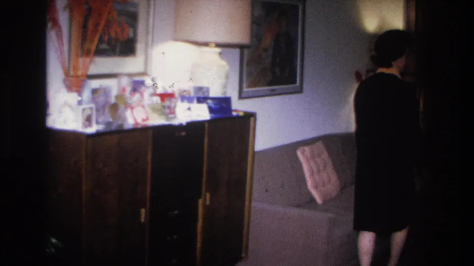 Woman in mid-1960s dress standing at a credenza in a home interior; small framed photos and decorative objects on sideboard top, framed wall prints above.