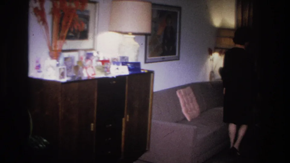 Woman in 1960s dark dress exiting frame right past a credenza in a mid-century living room; decorative objects and framed art on wall remain visible.