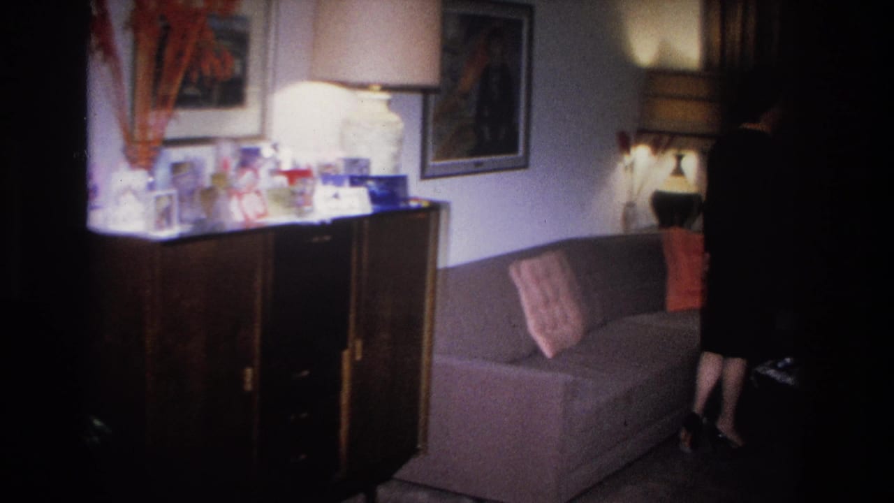 1966 Hoboken, NJ home movie: dark wood credenza with framed photos, drum-shade lamp, and wall art at left; grey sofa and woman in black dress partially entering frame at right.