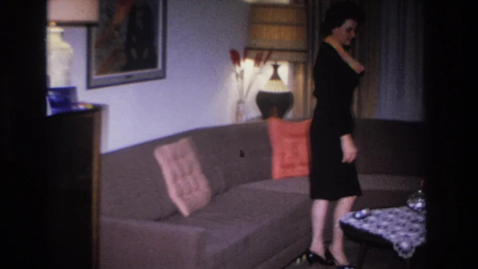 1966 Hoboken, NJ home movie: woman in fitted black dress and dark heels standing next to a grey mid-century sofa; credenza, ceramic lamp, and wall art behind her.