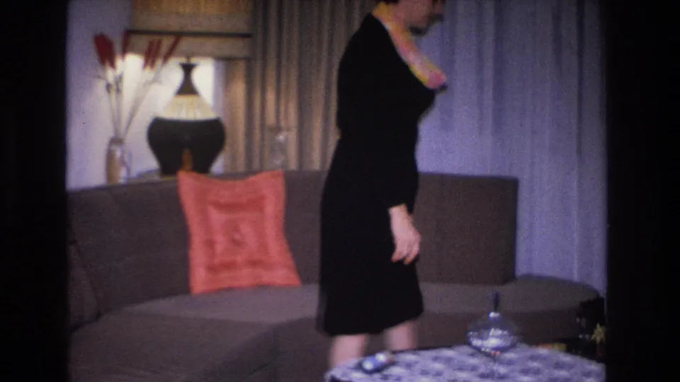 1966 Hoboken, NJ home movie: woman in black dress with orange-yellow necklace or scarf accent leaning forward near the sofa; ceramic lamp and floor-length drapes in background.