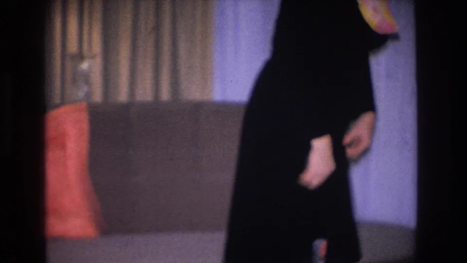 1966 Hoboken, NJ home movie: tight crop on woman in fitted black dress with orange-yellow necklace or scarf accent; salmon cushion and curtains visible in background.