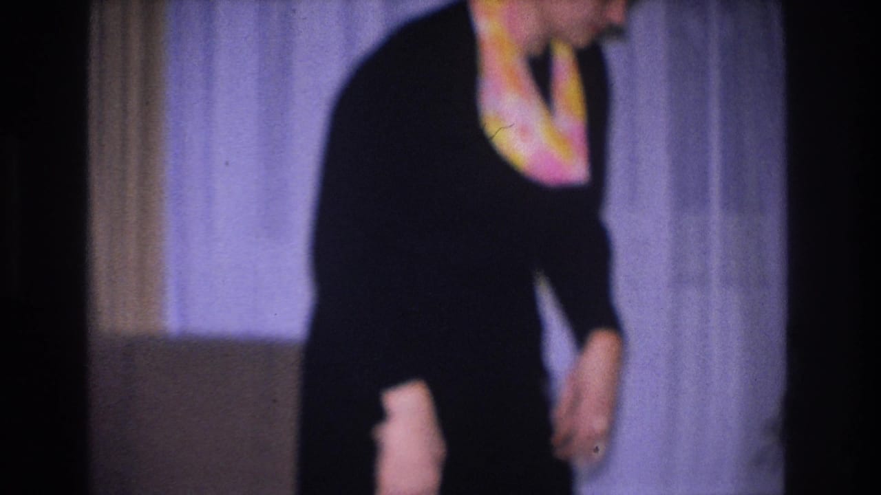 Close crop of a woman in a black dress and colorful scarf looking downward inside a home, from an 8mm film shot in Hoboken, New Jersey, 1966.
