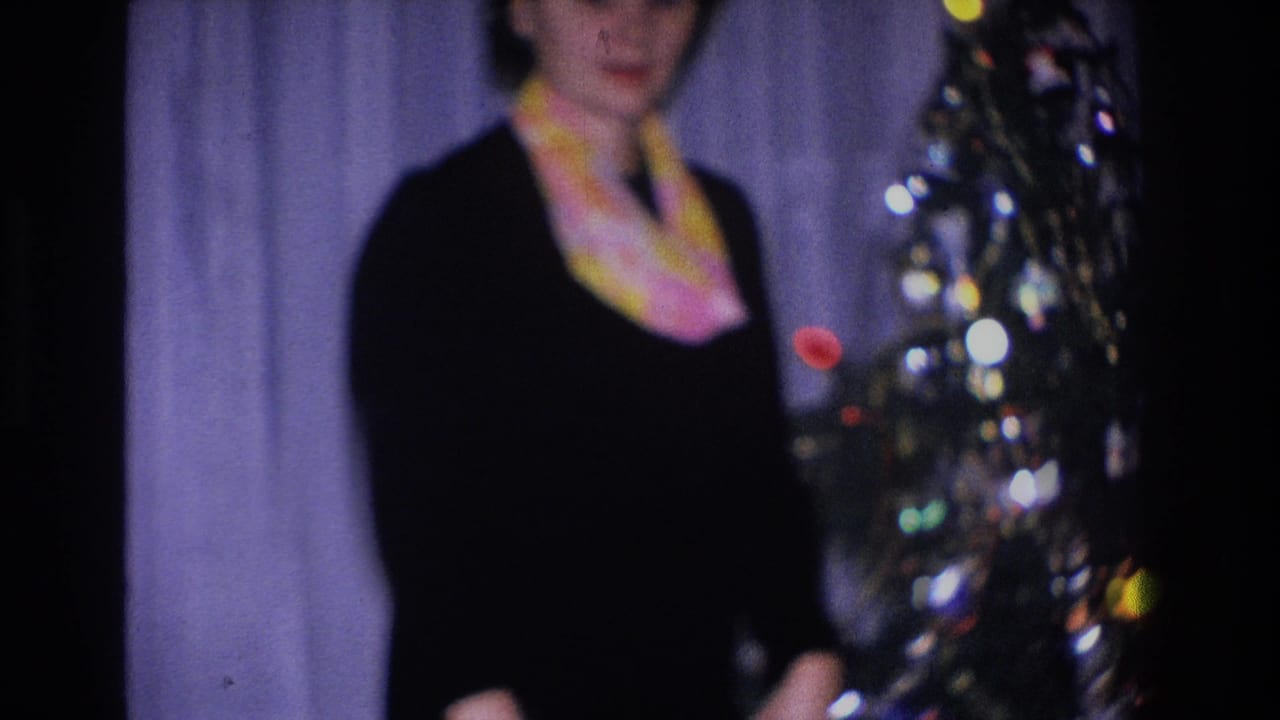 Woman in black dress and colorful scarf standing indoors, with edge of a Christmas tree showing multicolored lights at right.