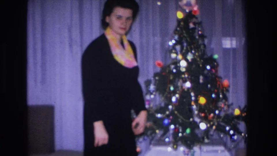 Woman in a black dress and colorful scarf posing beside a decorated Christmas tree with large multicolored bulbs and ornaments, in a private home interior, Hoboken 1966.