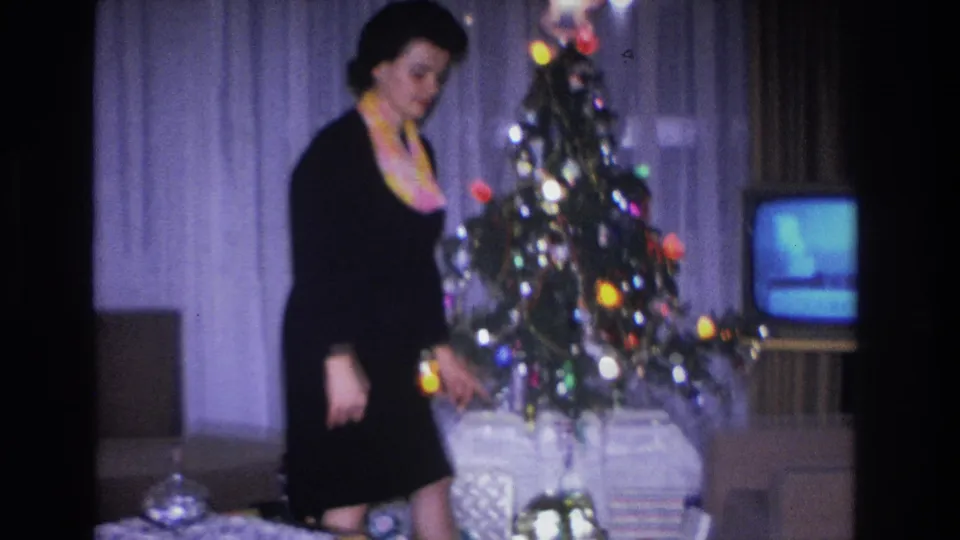 Woman bending toward wrapped gifts at the base of a decorated Christmas tree with multicolored lights; a small vintage CRT television is visible in the background, in a circa 1966 home interior.