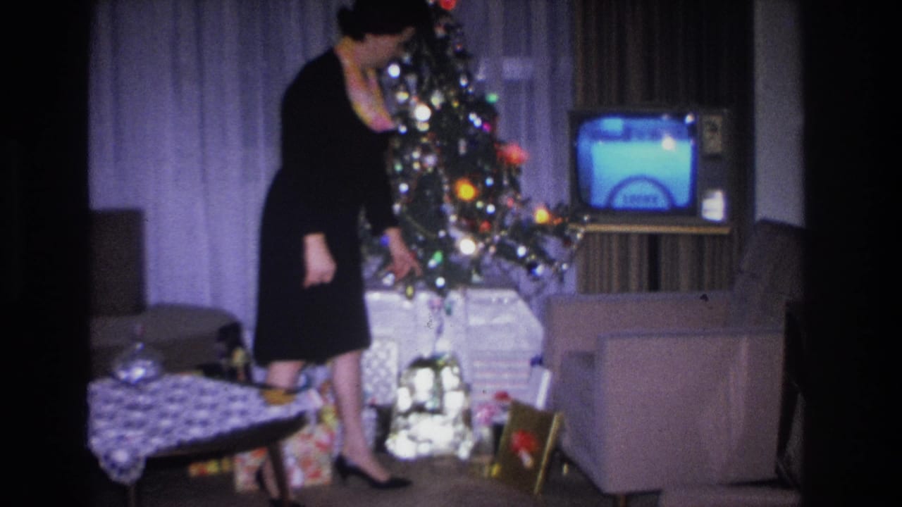 Woman in black dress standing beside a lit, ornamented Christmas tree with wrapped gifts below; small boxy television visible in background.