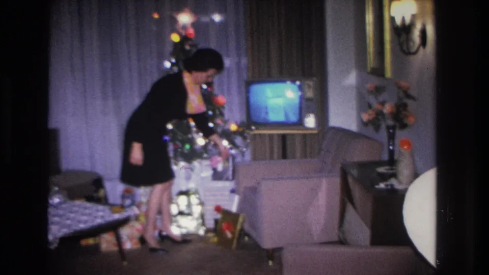 Woman in black dress crouching by a decorated Christmas tree in a mid-1960s living room, with a wall sconce lamp and flower arrangement visible in the background.