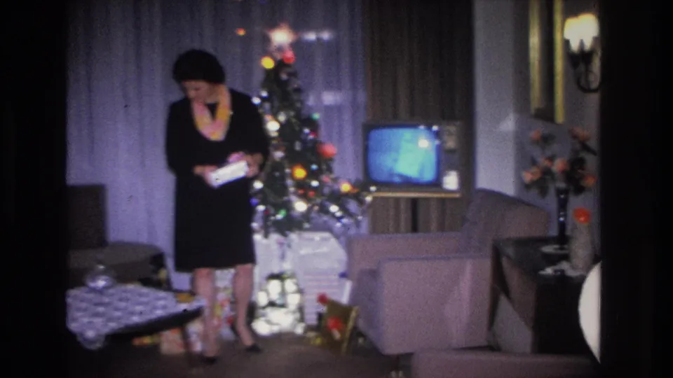 Woman in black dress standing before a lit Christmas tree in a mid-1960s living room, examining a gift or card; a second partially visible figure appears at the right edge of frame.