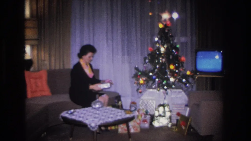 Woman in black dress and colorful scarf sits on a mid-century sofa examining a gift, with a lit Christmas tree and glowing vintage television in the background, c.1966 Hoboken living room.