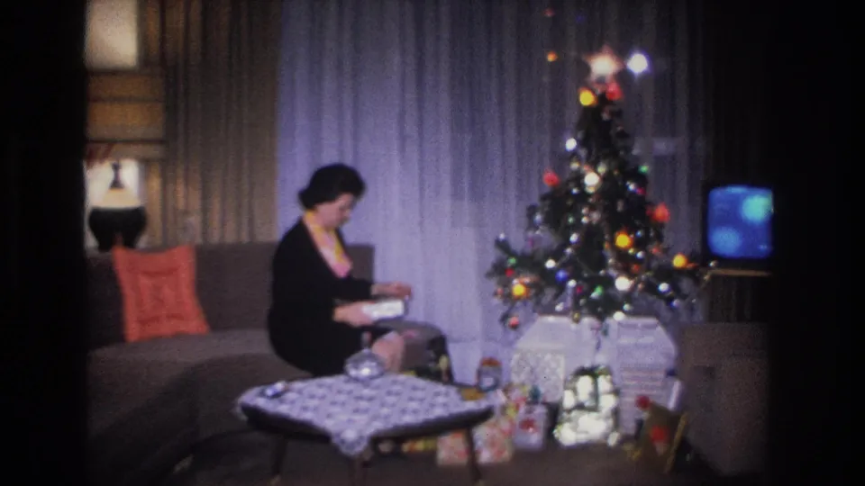 Woman in black dress and colorful scarf seated on a mid-century sofa holding an open gift, with a lit Christmas tree at center and vintage television at right, c.1966 Hoboken living room.