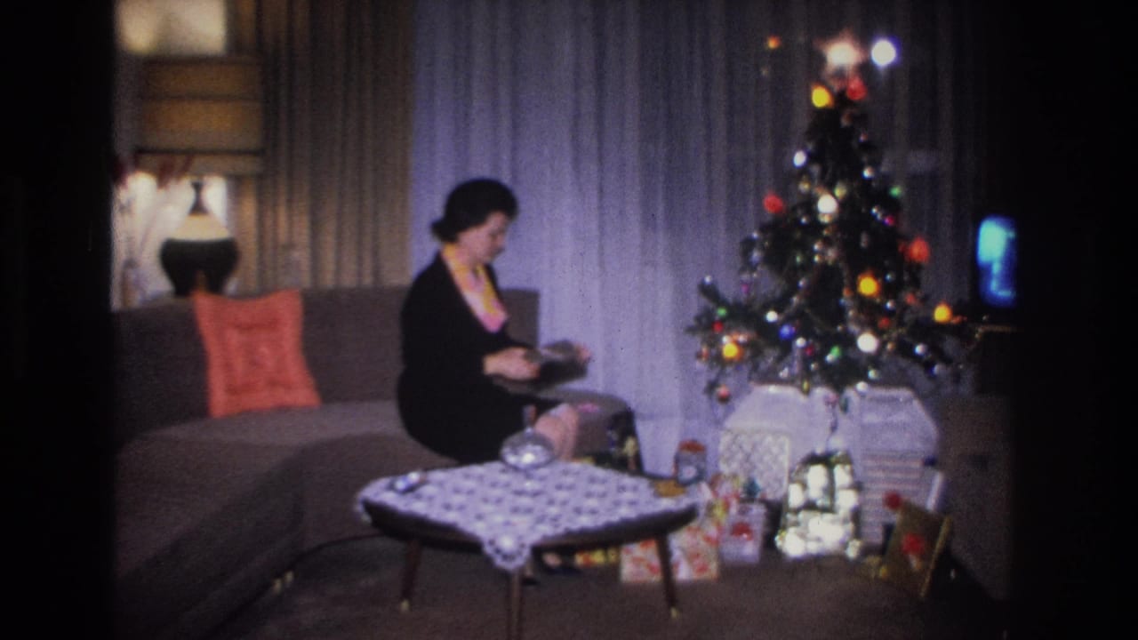 Woman in black dress opens a gift on a gray sofa beside a multicolored-bulb Christmas tree; wicker lamp, patterned coffee table, and blue-screen TV visible; Hoboken, NJ, c.1966.