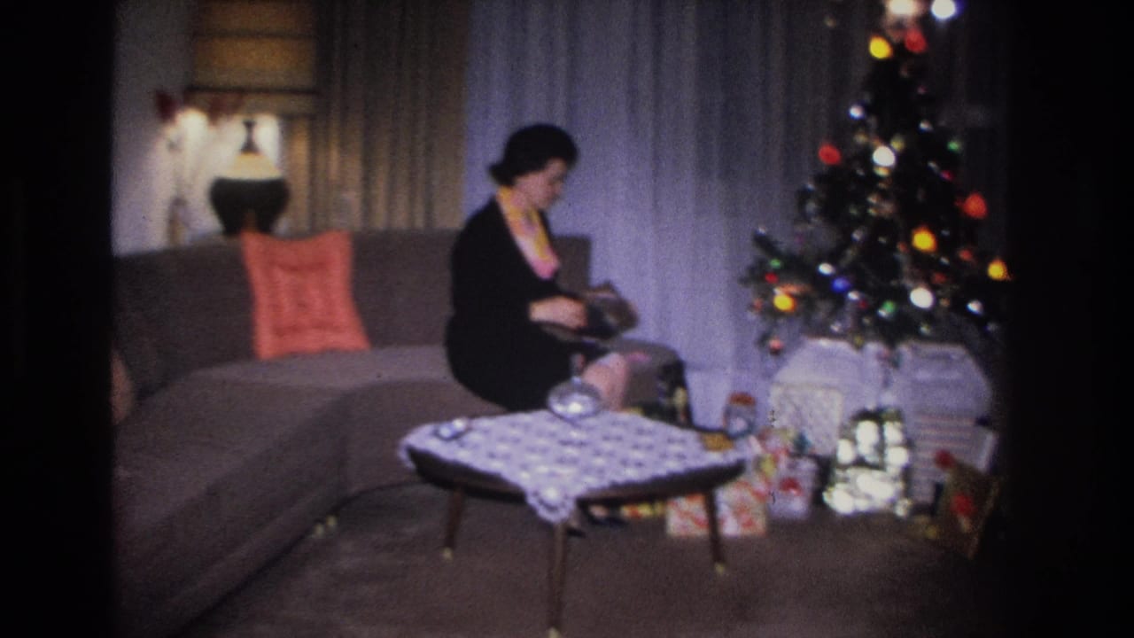 Woman on gray sofa examines an opened gift; Christmas tree with colored lights and wrapped presents visible to her right.