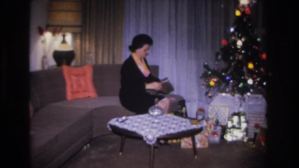 Woman in black dress with colorful necklace inspects gift contents on a gray sofa; multicolored Christmas tree lights and wrapped floor presents behind her; Hoboken, NJ, c.1966.
