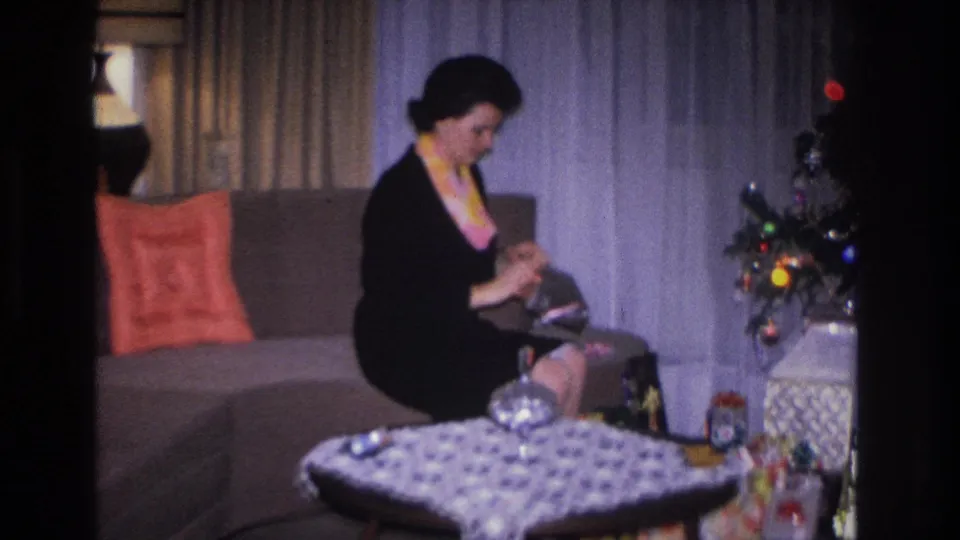 Woman in black dress with colorful necklace handles a gift on her lap; red throw pillow, patterned coffee table, and Christmas tree edge visible; Hoboken, NJ, c.1966.