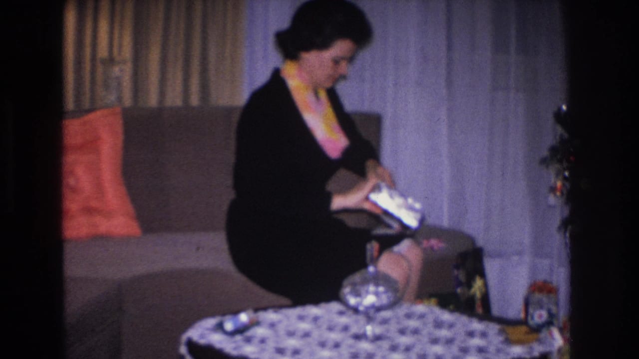 Woman on upholstered sofa handling a gift box in a 1960s living room; sheer curtains, patterned tablecloth, and orange cushion visible; circa 1966 8mm home movie, Hoboken NJ.