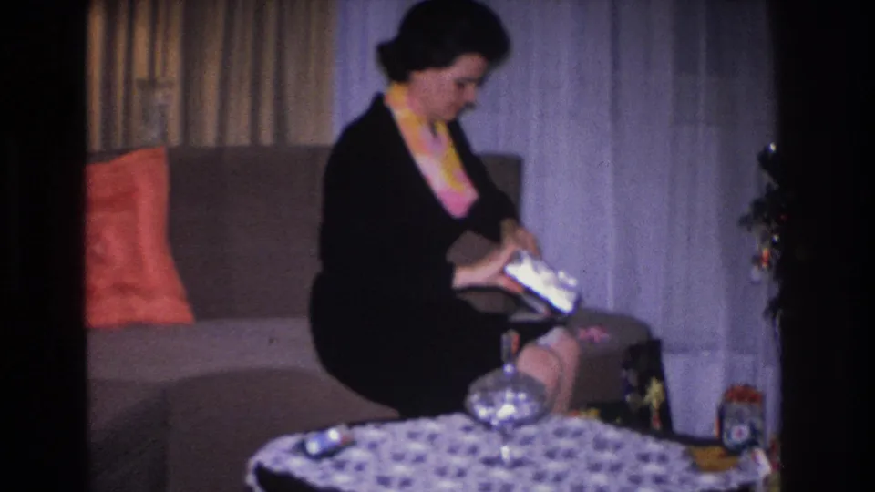 Woman on upholstered sofa handling a gift box in a 1960s living room; sheer curtains, patterned tablecloth, and orange cushion visible; circa 1966 8mm home movie, Hoboken NJ.
