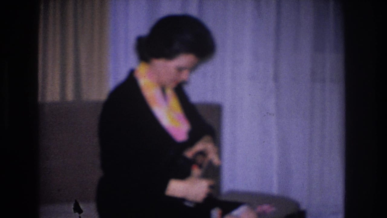 Woman in black dress and yellow-pink scarf examines a small object in her hands; 1966 8mm home movie, Hoboken, New Jersey.