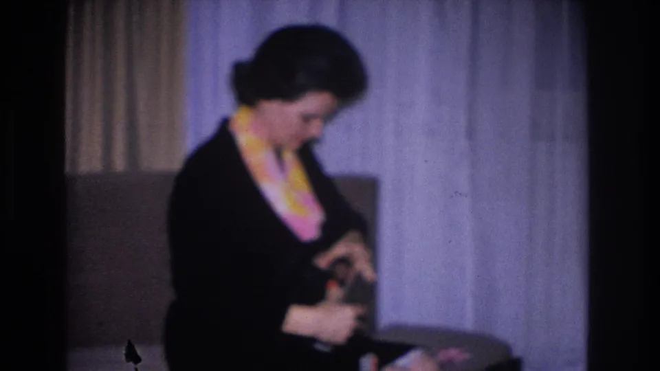 Woman in black dress and yellow-pink scarf examines a small object in her hands; 1966 8mm home movie, Hoboken, New Jersey.