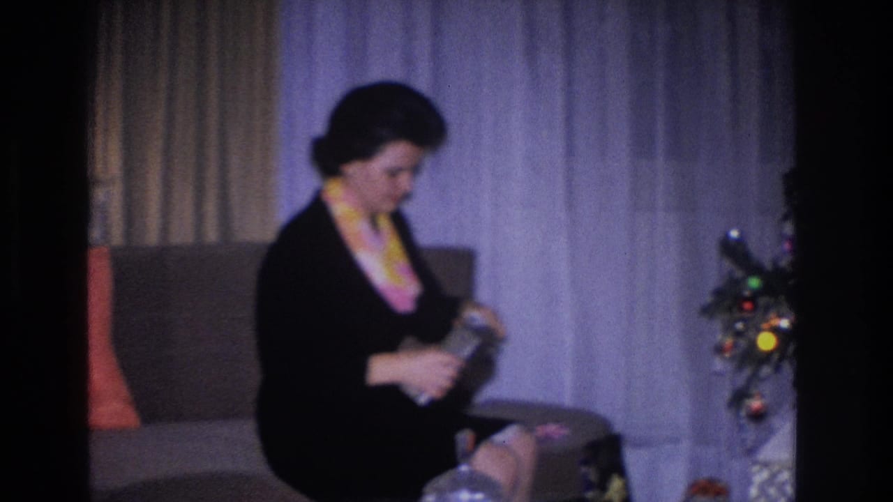 Woman in black dress seated on tan sofa with orange cushion; decorated Christmas tree with multicolor bulb lights visible at right edge.