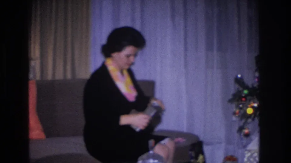 Woman in black dress and yellow-pink scarf on upholstered sofa; Christmas tree with multicolor incandescent bulb lights at right; 1966 8mm home movie, Hoboken, NJ.