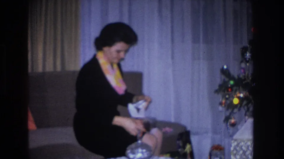 Woman in black dress and yellow-pink scarf unwraps a gift box in a domestic living room; decorated Christmas tree with multicolor incandescent lights at right; 1966, Hoboken, NJ.