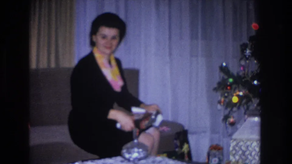 Woman in black dress and yellow-pink scarf smiles holding an open gift box; Christmas tree with large multicolor incandescent bulb lights and wrapped gifts at right; 1966 home movie, Hoboken, NJ.