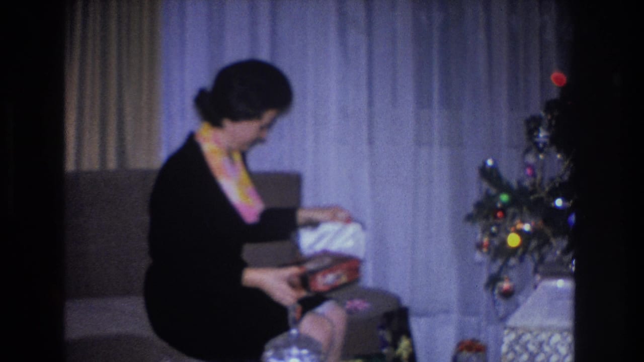 Mid-1960s woman in black dress and yellow scarf unwrapping a gift on an upholstered couch, small ornament-laden Christmas tree visible in background; Hoboken, NJ, circa 1966.