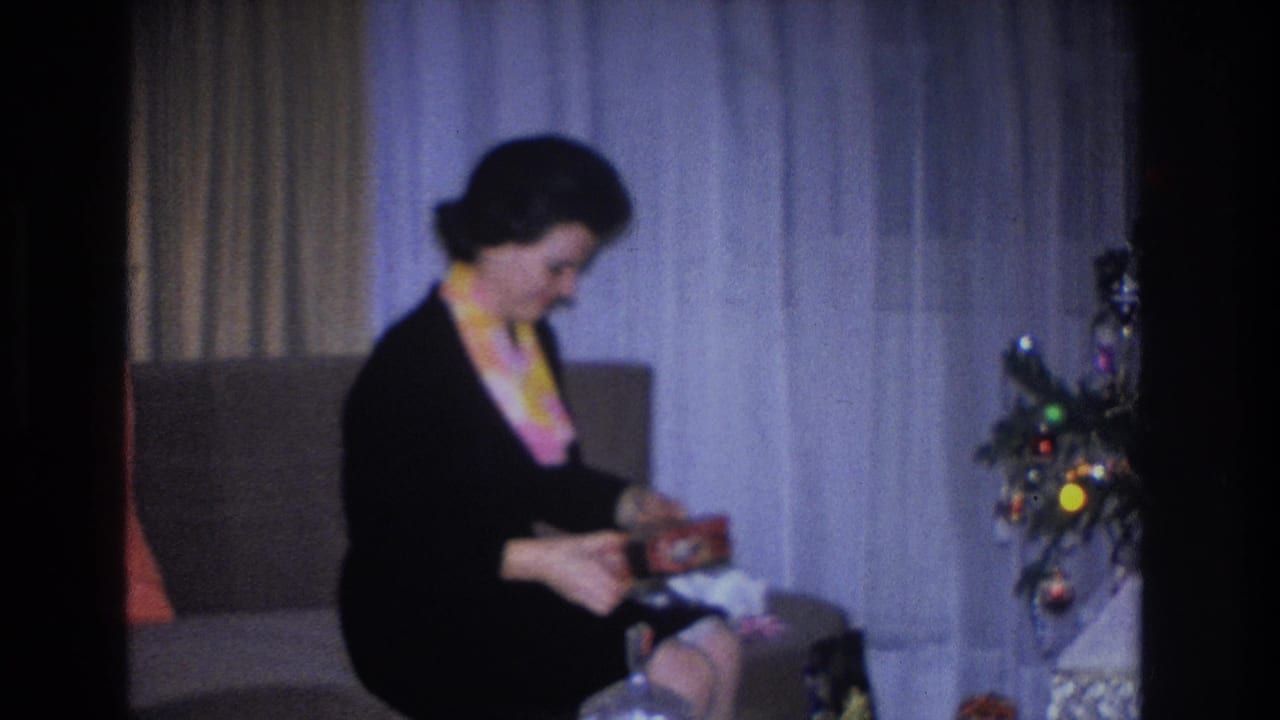 Woman in black dress leaning forward to examine contents of an opened gift box, Christmas tree with colored ornaments at right.