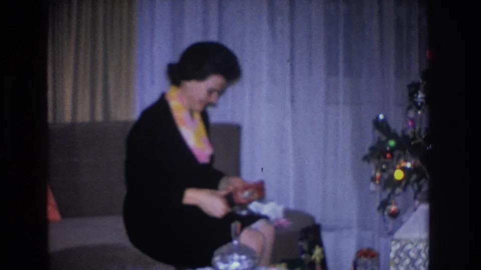 Woman in black dress leaning over an unwrapped gift box with tissue paper emerging; small Christmas tree with multicolored ornaments at right; domestic interior, circa 1966, Hoboken, NJ.